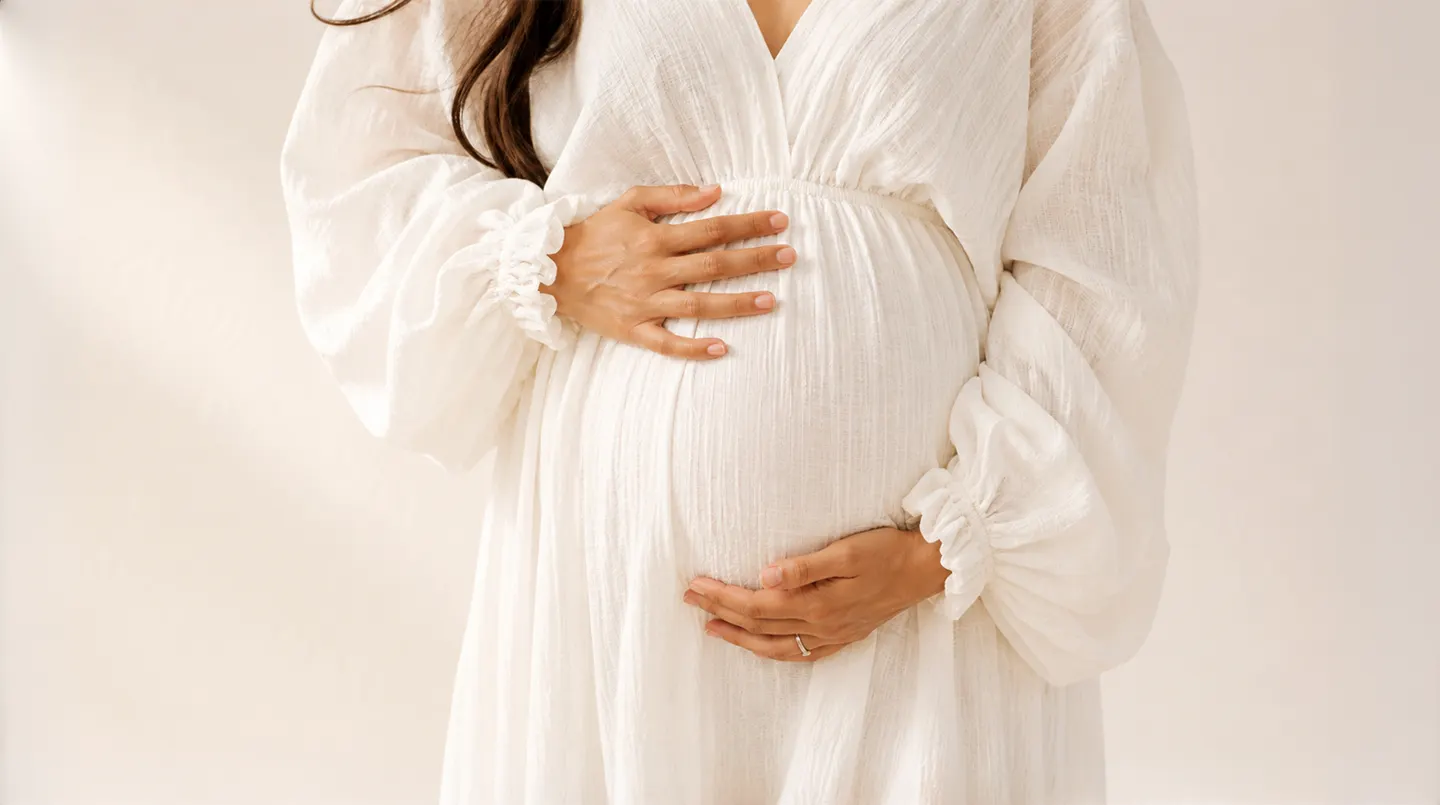 Schwangere Frau in einem weissen Kleid, die ihren Bauch mit beiden Händen hält